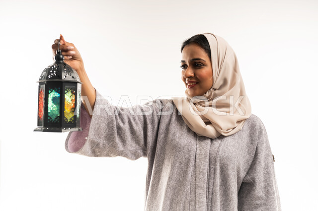 الأعياد والمناسبات الاسلامية, شابة سعودية تنظر إلى المصباح بإيماءات السرور, بورتريه مقرب لامرأة عربية خليجية عمانية محجبة ترتدي العباءة تمسك بيديها فانوس ملون, الاستعداد لاستقبال شهر رمضان المبارك, خلفية بيضاء