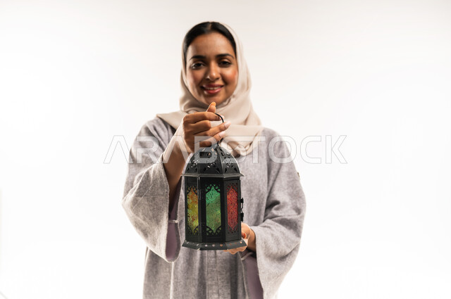 فوانيس وزينة الأعياد والمناسبات الاسلامية, شابة سعودية تنظر إلى الكاميرا بإيماءات السرور, بورتريه مقرب لامرأة عربية خليجية عمانية محجبة ترتدي العباءة تمسك بيديها فانوس ملون, الاستعداد لاستقبال شهر رمضان المبارك , خلفية بيضاء