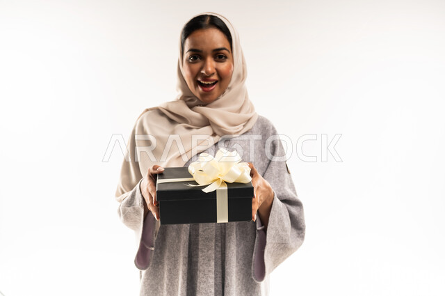 التعبير عن المحبة والتقدير, تقديم الهدايا في المناسبات السعيدة,  الاحتفال بأعياد الميلاد, شابة سعودية تنظر إلى الكاميرا بإيماءات المفاجاة والسرور, بورتريه مقرب لامرأة عربية خليجية عمانية محجبة ترتدي العباءة تمسك بيديها علبة هداية, خلفية بيضاء