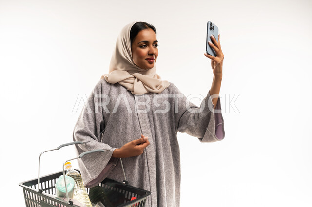 الاستمتاع بالتسوق في المولات والمراكز التجارية، شابة عمانية تقوم بشراء المستلزمات والحاجيات الضرورية، الاستفادة من  العروض والخصومات، بورتريه لامرأة عربية خليجية سعودية محجبة ترتدي العباءة تحمل بيدها السلة، شراء المنتجات من السوبر ماركت، خلفية بيضاء