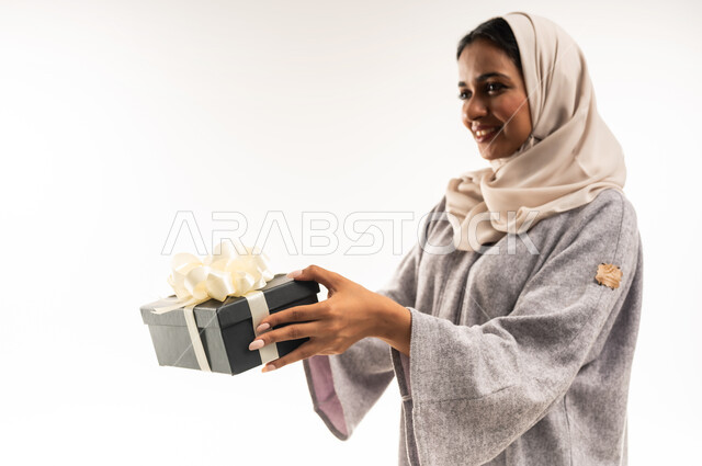 الاحتفال بأعياد الميلاد, التعبير عن المحبة والتقدير, شابة سعودية تنظر إلى مكان ما بإيماءات السعادة والسرور, تقديم الهدايا في المناسبات السعيدة, بورتريه مقرب من الجانب لامرأة عربية خليجية عمانية محجبة ترتدي العباءة تمسك بيديها علبة هداية, خلفية بيضاء