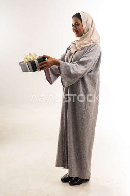 التعبير عن المحبة والتقدير, شابة سعودية تنظر إلى مكان ما بإيماءات السعادة والسرور, تقديم الهدايا في المناسبات السعيدة, الاحتفال بأعياد الميلاد, بورتريه من الجانب لامرأة عربية خليجية عمانية محجبة ترتدي العباءة تمسك بيديها علبة هداية, الجسم بالطول الكامل، خلفية بيضاء