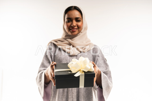 شابة سعودية تنظر إلى الكاميرا بإيماءات السعادة والسرور, تقديم الهدايا في المناسبات السعيدة, الاحتفال بأعياد الميلاد, التعبير عن المحبة والتقدير, بورتريه مقرب لامرأة عربية خليجية عمانية محجبة ترتدي العباءة تمسك بيديها علبة هداية, خلفية بيضاء