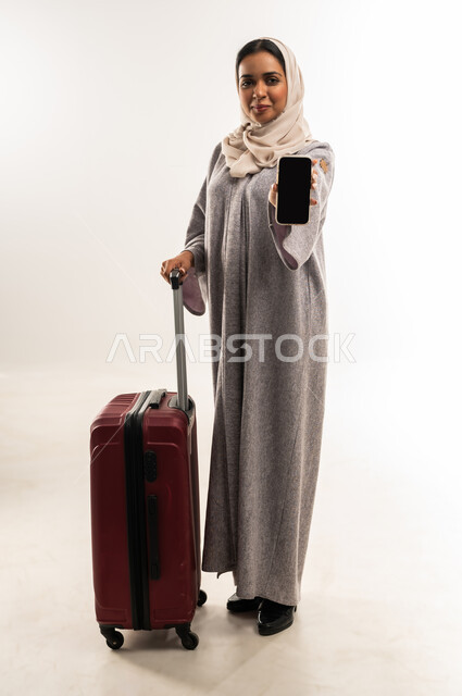 عرض شاشة سوداء عبر الهاتف المحمول، الشعور بالسعادة والفرح، السياحة في إجازة الصيف, شابة سعودية تستعد لقضاء رحلة صيفية ممتعة, بورتريه لشابة عربية خليجية عمانية محجبة ترتدي العباءة تمسك حقيبة السفر, استخدم الأجهزة والتقنيات الحديثة والمتطورة،خلفية بيضاء