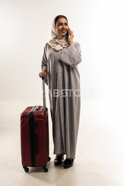 الإجابة والرد على الاتصالات باستخدام الهاتف المحمول، الشعور بالسعادة والفرح، السياحة في إجازة الصيف, شابة سعودية تستعد لقضاء رحلة صيفية ممتعة, بورتريه لشابة عربية خليجية عمانية محجبة ترتدي العباءة تمسك حقيبة السفر, التواصل والاطمئنان على الأصدقاء، خلفية بيضاء