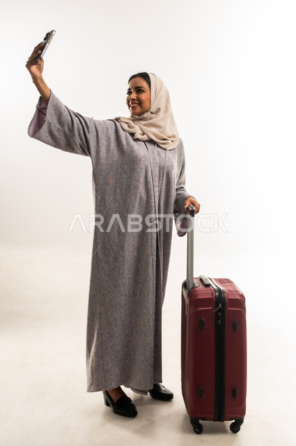 توثيق اللحظات التذكارية عبر جهاز الجوال، الشعور بالسعادة والفرح، السياحة في إجازة الصيف, شابة سعودية تستعد لقضاء رحلة صيفية ممتعة, بورتريه لشابة عربية خليجية عمانية محجبة ترتدي العباءة تمسك حقيبة السفر تقوم بالتقاط صورة سيلفي, الجسم بالطول الكامل, خلفية بيضاء