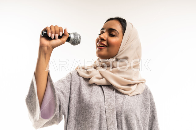 تقديم العروض الفنيه و الغنائيه والفقرات الترفيهية، سيدة عمانية تقوم بإلقاء الخطابات، مفهوم الفن وتأثيره الايجابي على الامرأة، بدء التدريب الصوتي، بورتريه مقرب لشابة عربية خليجية سعودية ترتدي العباءة تمسك بيدها المايكروفون بإيماءات الاندماج بالغناء، خلفيه بيضاء