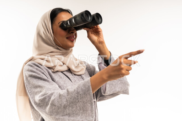 رؤية الأشياء البعيدة، التفكير والتطلع إلى المستقبل، التخطيط المستقبلي للأهداف والطموحات، سيدة سعودية تشير باصبع السبابة إلى شيء ما، بورتريه مقرب لشابة عربية خليجية عمانية ترتدي العباءة والحجاب تمسك منظار ثنائي العينين تنظر إلى شيء ما، خلفية بيضاء