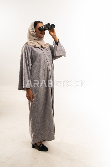 التخطيط المستقبلي للأهداف والطموحات، سيدة سعودية تقوم برؤية الأشياء البعيدة، حب الاستكشاف والمغامرة، بورتريه لشابة عربية خليجية عمانية ترتدي العباءة والحجاب تمسك منظار ثنائي العينين تنظر إلى شيء ما، التفكير والتطلع إلى المستقبل، الجسم بالطول الكامل، خلفية بيضاء