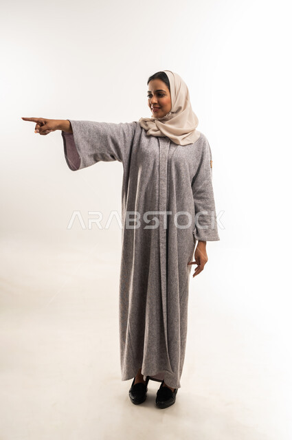 إشارات تدل على هذا المكان (هنا)، سيدة سعودية تقوم بتحديد موقع معين بإيماءات البشاشة، بورتريه لامرأة عربية خليجية عمانية مبتسمة ترتدي الحجاب والعباءة تنظر إلى الكاميرا ترفع أصبع السبابة للأعلى وتشير إلى شيء ما، الجسم بالطول الكامل، خلفية بيضاء
