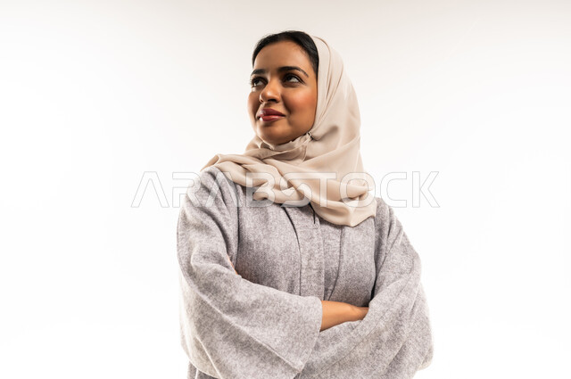 أزياء محجبات عصرية راقية، الملامح العربية الأصيلة، مفهوم الثقة بالنفس والاهتمام بالمظهر الخارجي، بورتريه لامرأة عربية خليجية سعودية مبتسمة ترتدي الحجاب والعباءة تنظر الى الكاميرا بتعابير السرور، شابة عمانية تقف بأيدي متقاطعة، خلفية بيضاء