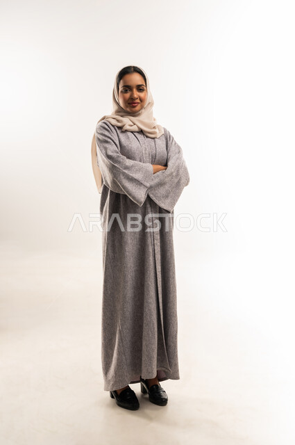 أزياء محجبات عصرية راقية، الملامح العربية الأصيلة، مفهوم الثقة بالنفس والاهتمام بالمظهر الخارجي، بورتريه لامرأة عربية خليجية سعودية مبتسمة ترتدي الحجاب والعباءة تنظر الى الكاميرا بتعابير السرور، شابة عمانية تقف بأيدي متقاطعة، الجسم بالطول الكامل، خلفية بيضاء
