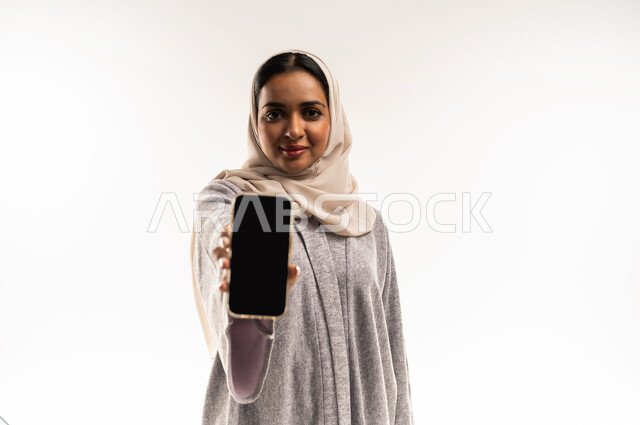 النظر الى الكاميرا بايماءات السعادة والسرور، بورتريه مقرب لإمرأة عربية خليجية عمانية مبتسمة ترتدي العباءة تقوم بعرض شاشة سوداء فارغة عبر الهاتف المحمول، شابة سعودية تستخدم الأجهزة والتقنيات الحديثة والمتطورة، خلفية بيضاء