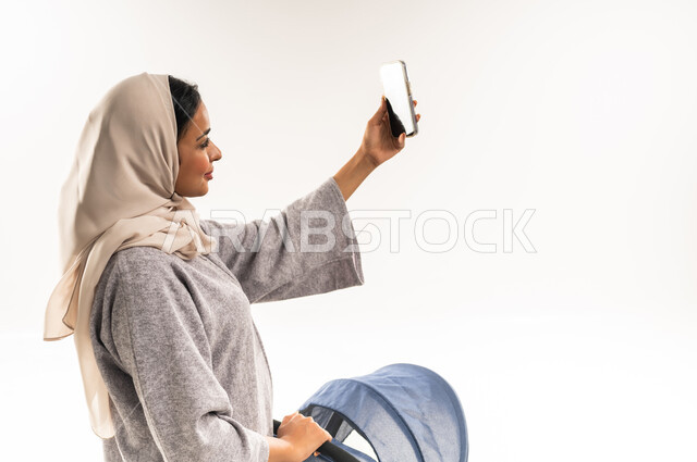 توثيق اللحظات التذكارية عبر جهاز الجوال, مفهوم الأمومة والرعايه والاهتمام بالأولاد، بورتريه لأم عربية خليجيه سعودية محجبة تقوم بجر عربة الأطفال, امراة عمانية تلتقط صورة سيلفي عبر الهاتف المحمول، القيام بالنزهات والجولات العائلية، خلفية بيضاء