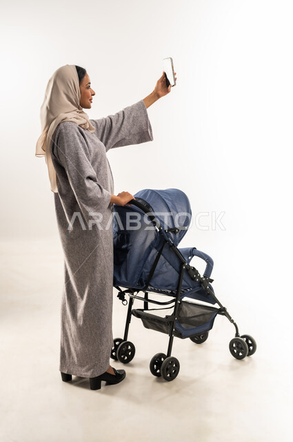 توثيق اللحظات التذكارية عبر جهاز الجوال, مفهوم الأمومة والرعايه والاهتمام بالأولاد، بورتريه لأم عربية خليجيه سعودية محجبة تقوم بجر عربة الأطفال, امراة عمانية تلتقط صورة سيلفي عبر الهاتف المحمول، القيام بالنزهات والجولات العائلية، خلفية بيضاء