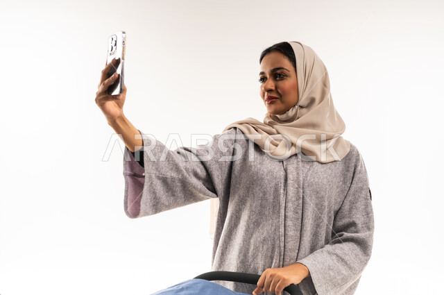 توثيق اللحظات التذكارية عبر جهاز الجوال, مفهوم الأمومة والرعايه والاهتمام بالأولاد، بورتريه لأم عربية خليجيه سعودية محجبة تقوم بجر عربة الأطفال, امراة عمانية تلتقط صورة سيلفي عبر الهاتف المحمول، القيام بالنزهات والجولات العائلية، خلفية بيضاء