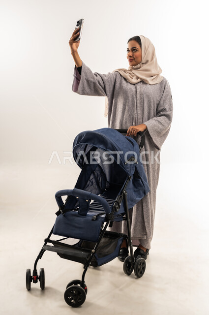 توثيق اللحظات التذكارية عبر جهاز الجوال, مفهوم الأمومة والرعايه والاهتمام بالأولاد، بورتريه لأم عربية خليجيه سعودية محجبة تقوم بجر عربة الأطفال, امراة عمانية تلتقط صورة سيلفي عبر الهاتف المحمول، القيام بالنزهات والجولات العائلية، خلفية بيضاء