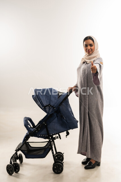أهمية الأم ودورها الأساسي في تربية ونشأة الأبناء, مفهوم الأمومة والرعايه والاهتمام بالأولاد، بورتريه لأم عربية خليجيه سعودية محجبة تقوم بجر عربة الأطفال, امراة عمانية ترفع اصبع الابهام بايماءات الجودة والامتياز، القيام بالنزهات والجولات العائلية، خلفية بيضاء
