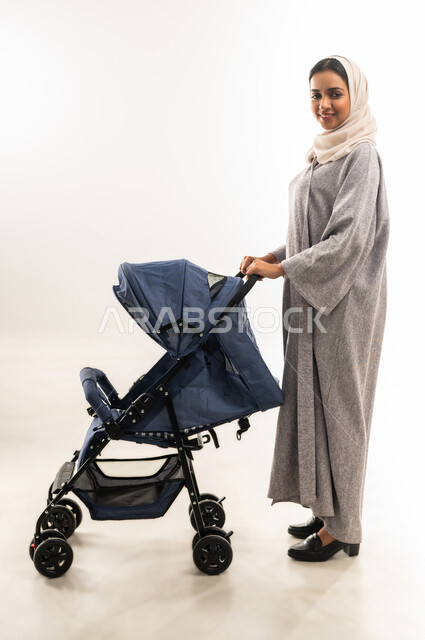 أهمية الأم ودورها الأساسي في تربية ونشأة الأبناء, القيام بالنزهات والجولات العائلية، مفهوم الأمومة والرعايه والاهتمام بالأولاد،  بورتريه لأم عربية خليجيه سعودية محجبة تقوم بجر عربة الأطفال, امراة عمانية تنظر إلى الكاميرا بايماءات السعادة والسرور، خلفية بيضاء