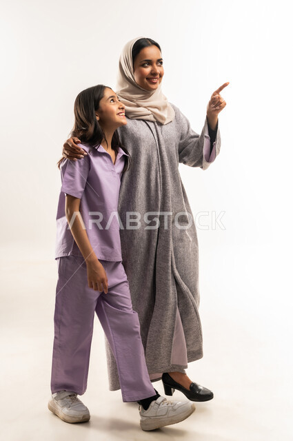 الاشارة باصبع السبابة إلى شيء ما، عائلة عمانية تقضي اوقات ممتعة، النظر الى مكان ما بايماءات السعادة والسرور، بورتريه لامرأة عربية خليجية سعودية محجبة ترتدي عباءة ملونة تقف مع ابنتها، تبادل الحوارات والنقاشات المسلية، تعزيز العلاقات الاجتماعية، خلفية بيضاء
