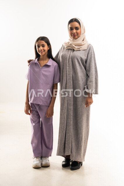 النظر الى الكاميرا بايماءات السعادة والسرور، بورتريه لامرأة عربية خليجية سعودية محجبة ترتدي عباءة ملونة تقف مع ابنتها، عائلة عمانية تقضي اوقات ممتعة، تبادل الحوارات والنقاشات المسلية، تعزيز العلاقات الاجتماعية، الجسم بالطول الكامل، خلفية بيضاء