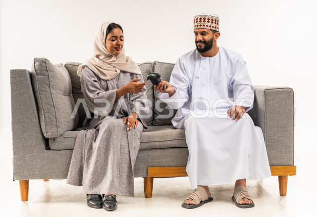 إيماءات السرور والتحدي والمتعة، الاستمتاع والتسلية في العطلة، مفهوم الاوقات العائلية السعيده، بورتريه لزوج عربي خليجي عماني يرتدي الدشداشة والكمة يقوم بإعطاء عصا التحكم لزوجته، الجلوس على كنبه مريحه واللعب بألعاب الفيديو، خلفيه بيضاء