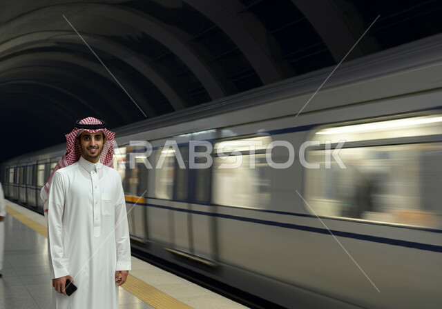 النظر إلى الكاميرا بإيماءات السعادة والسرور, الاستعداد لركوب القطار, توفير وتسهيل خدمات النقل الحديثة, تطور وتقدم وسائل المواصلات في المملكة, رجل عربي خليجي سعودي يرتدي الثوب التقليدي والشماغ يقف في محطة مترو الرياض, مفهوم التنقل السريع والآمن في السعودية