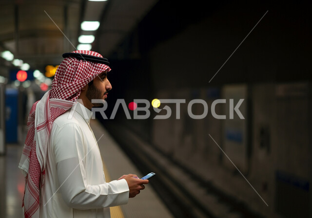 وسائل النقل والمواصلات البرية الحديثة في المملكة, تسهيل الحركة المرورية في السعودية, استخدام الأجهزة التقنية المتطورة, صورة مقربة من الجانب لرجل عربي خليجي سعودي يرتدي الثوب التقليدي والشماغ يقف في محطة مترو الرياض ويمسك الهاتف المحمول بيده