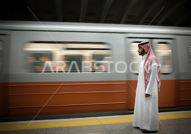 الاستعداد لركوب القطار, توفير وتسهيل خدمات النقل الحديثة, تطور وتقدم وسائل المواصلات في المملكة, صورة من الجانب لرجل عربي خليجي سعودي يرتدي الثوب التقليدي والشماغ يقف في محطة مترو الرياض, مفهوم التنقل السريع والآمن في السعودية