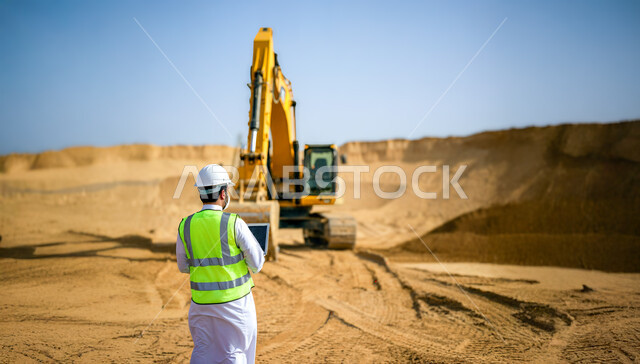 دمج التقنيات الحديثة بأعمال التعدين, صورة من الخلف لمهندس عربي خليجي سعودي يرتدي سترة وخوذة الحماية يستعمل الحاسوب المحمول, التنقيب عن الثروات الباطنية في المناطق الصحراوية, جرافة تقوم بالتخلص من الأتربة والصخور, التشكيلات والتكوينات الصخرية في صحاري السعودية