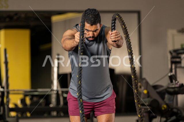 ايماءات القوة والعزم، أسلوب حياة صحي، مفهوم الرشاقة واللياقة البدنية، المحافظة على توازن الجسم، انشطة ورياضات لتقوية العضلات والاعصاب، رجل عربي خليجي اماراتي يرتدي الملابس الرياضية يقوم بممارسة التمارين بواسطة الحبل الثقيل، شاب سعودي يتمرن داخل النادي الرياضي