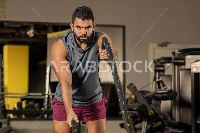 ايماءات القوة والعزم، أسلوب حياة صحي، مفهوم الرشاقة واللياقة البدنية، المحافظة على توازن الجسم، انشطة ورياضات لتقوية العضلات والاعصاب، رجل عربي خليجي اماراتي يرتدي الملابس الرياضية يقوم بممارسة التمارين بواسطة الحبل الثقيل، شاب سعودي يتمرن داخل النادي الرياضي