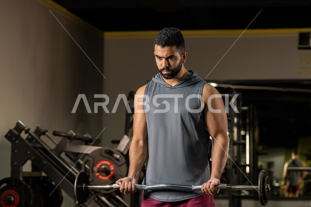 ايماءات القوة والعزم، الاهتمام بالصحة واللياقة الجسدية، رياضة كمال الأجسام، المحافظة على توازن الجسم، شاب عربي خليجي إماراتي يرتدي الزي الرياضي يقوم بممارسة تمارين رفع الأوزان، رجل سعودي يحمل بيده البار، أنشطة لتقوية العضلات، أسلوب حياة صحي