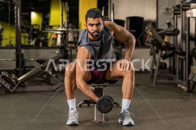 إيماءات القوة والعزم، الاهتمام بالصحة واللياقة الجسدية، رياضة كمال الأجسام، استخدام الاثقال الرياضية، شاب عربي خليجي اماراتي يرتدي الزي الرياضي يقوم بممارسة تمارين رفع الأوزان، رجل سعودي يحمل بيده الدامبلز، بناء الكتلة العضلية للجسم