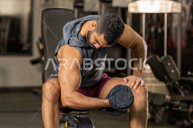 إيماءات القوة والعزم، الاهتمام بالصحة واللياقة الجسدية، رياضة كمال الأجسام، استخدام الاثقال الرياضية، شاب عربي خليجي اماراتي يرتدي الزي الرياضي يقوم بممارسة تمارين رفع الأوزان، رجل سعودي يحمل بيده الدامبلز، بناء الكتلة العضلية للجسم