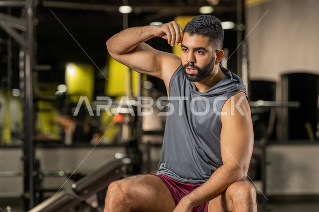 مفهوم الرشاقة واللياقة البدنية, النظر والتركيز في شيء ما، رجل عربي خليجي إماراتي يرتدي الملابس الرياضية يقوم بعرض عضلاته, أسلوب حياة صحي، المحافظة على توازن الجسم، شاب سعودي يتمرن داخل النادي الرياضي