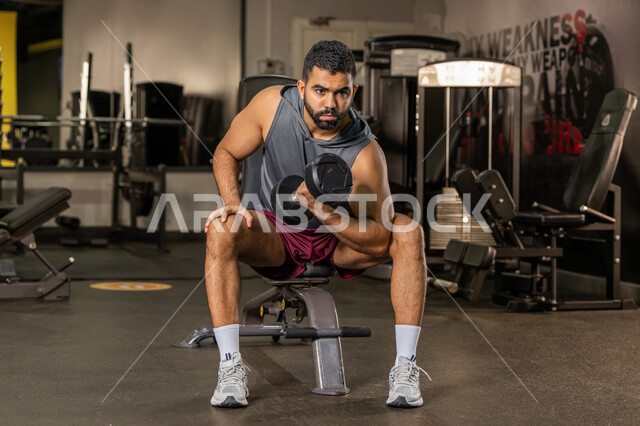 إيماءات القوة والعزم، الاهتمام بالصحة واللياقة الجسدية، رياضة كمال الأجسام، استخدام الاثقال الرياضية، شاب عربي خليجي اماراتي يرتدي الزي الرياضي يقوم بممارسة تمارين رفع الأوزان، رجل سعودي يحمل بيده الدامبلز، بناء الكتلة العضلية للجسم