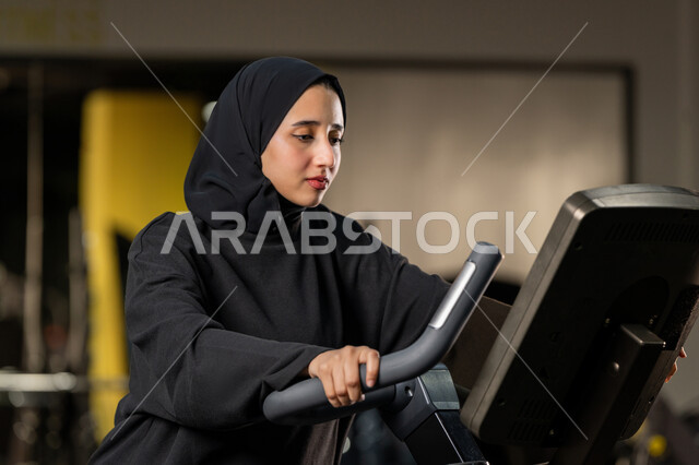 شابة عربية خليجية إماراتية محجبة ترتدي الزي الرياضي تقوم بممارسة التمارين على جهاز التجذيف، نشاط شبابي يساعد على اللياقة البدنية, أهمية الرياضة لبناء جسد قوي، حرق السعرات الحرارية وإنقاص الوزن، امراة سعودية تمارس تمرين لتحسين وضعية الجسم والتوازن