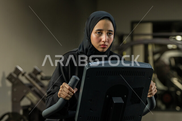 أهمية الرياضة لبناء جسد قوي، حرق السعرات الحرارية وإنقاص الوزن، نشاط شبابي يساعد على اللياقة البدنية, امراة سعودية تمارس تمرين لتحسين وضعية الجسم والتوازن، شابة عربية خليجية إماراتية محجبة ترتدي الزي الرياضي تقوم بممارسة التمارين على جهاز التجذيف