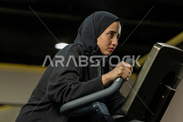 نشاط شبابي يساعد على اللياقة البدنية, أهمية الرياضة لبناء جسد قوي، حرق السعرات الحرارية وإنقاص الوزن، امراة سعودية تمارس تمرين لتحسين وضعية الجسم والتوازن، شابة عربية خليجية إماراتية محجبة ترتدي الزي الرياضي تقوم بممارسة التمارين على جهاز التجذيف