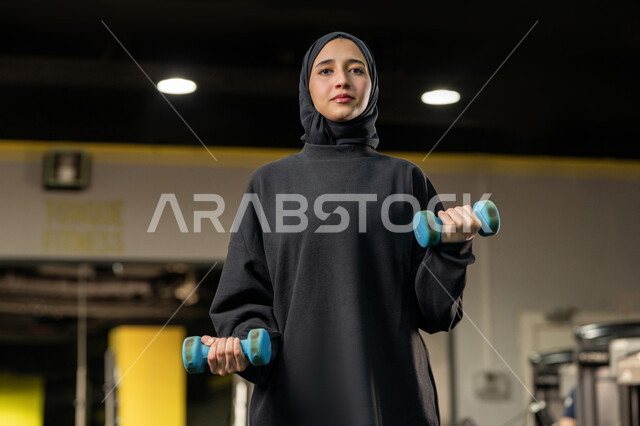 Burning fat and calories, preparing to exercise with gestures of enthusiasm and happiness, a young Emirati woman lifting weights, a sports activity to maintain a healthy body, a veiled Saudi Arabian Gulf woman wearing sportswear doing weightlifting exercises (dumbbells)