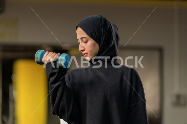 الاستعداد لممارسة التمارين الرياضية بايماءات الحماس والسعادة ، شابة اماراتية تقوم بحمل الأوزان، امراة عربية خليجية سعودية محجبة ترتدي الملابس الرياضة تقوم بتمارين رفع الأثقال (دمبلز)، نشاط رياضي للمحافظة على صحة الجسم، حرق الدهون والسعرات الحراريه