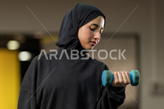 حرق الدهون والسعرات الحراريه، الاستعداد لممارسة التمارين الرياضية بايماءات الحماس والسعادة ، شابة اماراتية تقوم بحمل الأوزان، نشاط رياضي للمحافظة على صحة الجسم، امراة عربية خليجية سعودية محجبة ترتدي الملابس الرياضة تقوم بتمارين رفع الأثقال (دمبلز)