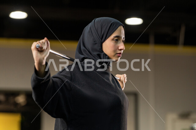 زيادة المرونة وتمديد العضلات، نشاط شبابي يساعد على اللياقة البدنية, أهمية الرياضة لبناء جسد قوي، امراة سعودية تمارس تمرين لتحسين وضعية الجسم والتوازن، شابة عربية خليجية إماراتية محجبة ترتدي الزي الرياضي تتمرن باستخدام العصا، أسلوب حياة صحي