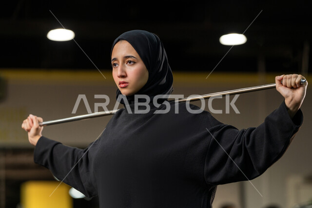 أسلوب حياة صحي، تمديد العضلات وزيادة المرونة، نشاط شبابي يساعد على اللياقة البدنية, أهمية الرياضة لبناء جسد قوي، امراة سعودية تمارس تمرين لتحسين وضعية الجسم والتوازن، شابة عربية خليجية إماراتية محجبة ترتدي الزي الرياضي تتمرن باستخدام العصا
