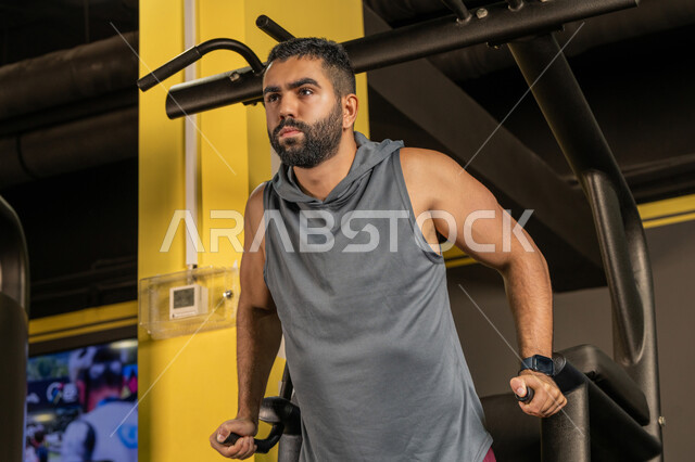 استخدام الأجهزة الرياضية، أسلوب حياة صحي، أنشطة شبابية تساعد على اللياقة البدنية, أهمية الرياضة لبناء جسد قوي، رجل سعودي يمارس تمرين لتقوية عضلات الصدر، رياضة كمال الأجسام، شاب عربي خليجي إماراتي يرتدي الزي الرياضي يقوم بممارسة التمارين على جهاز الغطس المساعد