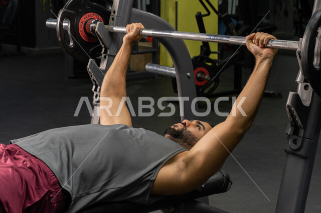 شاب عربي خليجي إماراتي يرتدي الزي الرياضي يقوم بممارسة تمارين رفع الأوزان، رجل سعودي يمارس أنشطة لتقوية العضلات، الاهتمام بالصحة واللياقة الجسدية، أسلوب حياة صحي، رياضة كمال الأجسام، إيماءات القوة والثقة، استخدام الأجهزة الرياضية