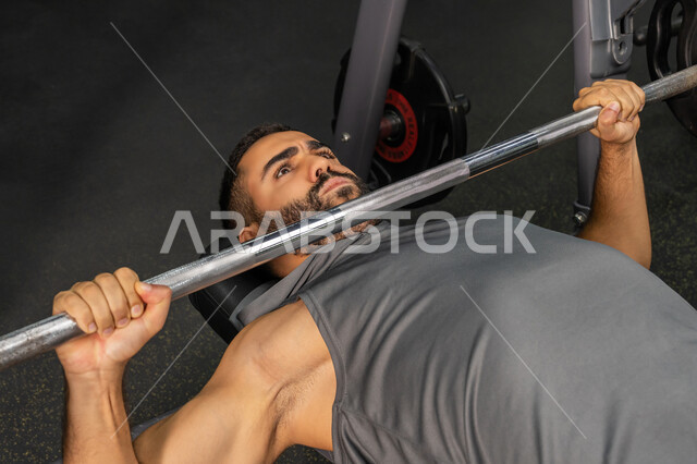 إيماءات القوة والثقة، رياضة كمال الأجسام، أسلوب حياة صحي، الاهتمام بالصحة واللياقة الجسدية، استخدام الأجهزة الرياضية، شاب عربي خليجي إماراتي يرتدي الزي الرياضي يقوم بممارسة تمارين رفع الأوزان، رجل سعودي يمارس أنشطة لتقوية العضلات