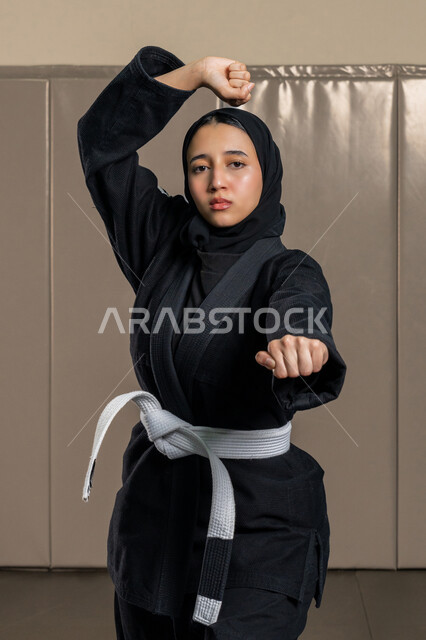 القيام بحركات قتالية خاصة برياضة الكاراتيه، شابة عربية خليجية سعودية محجبة ترتدي الزي الرياضي مع الحزام الأبيض تقوم بالتدرب على الفنون القتالية اليابانية, مفهوم اللياقة البدنية والتوازن، امراة اماراتية تمارس  رياضات وتمارين نسائية