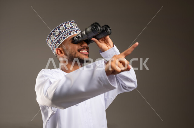 الإشارة بإصبع السبابة إلى شيء ما في الأعلى، حب الاستكشاف و المغامرة, بورتريه مقرب لشاب عربي خليجي عماني يرتدي الدشداشة و الكمة يحمل بيده منظار ثنائي العينين, مفهوم النظرة المستقبلية، رؤية الاشياء البعيدة عبر التلسكوب, خلفية بيج